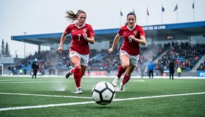 Calgary women’s soccer players sprint toward a ball during a match on a green turf field with a blurred crowd behind them.